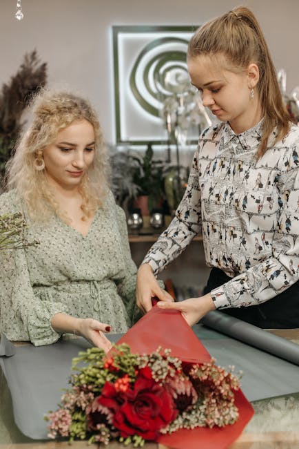 A florist from hackneymarshesflowers.co.uk is carefully wrapping a vibrant floral gift, featuring red roses, pink carnations, and sprigs of baby's breath, into red wrapping paper. The bouquet is posit