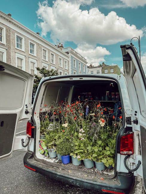 The open back of a white delivery van parked on a street shows an array of freshly cut flowers and greenery, including red poppies, white blossoms, and green foliage, arranged in blue and white pots. 