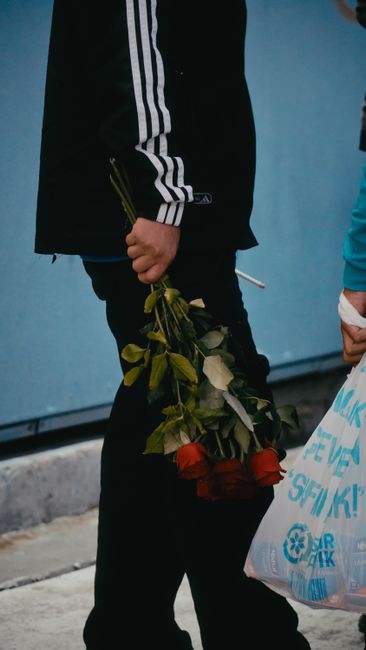 A person holding a bouquet of fresh flowers, including red and white roses with green foliage, wrapped in clear plastic. The individual is dressed in dark clothing with white stripe details on the sle