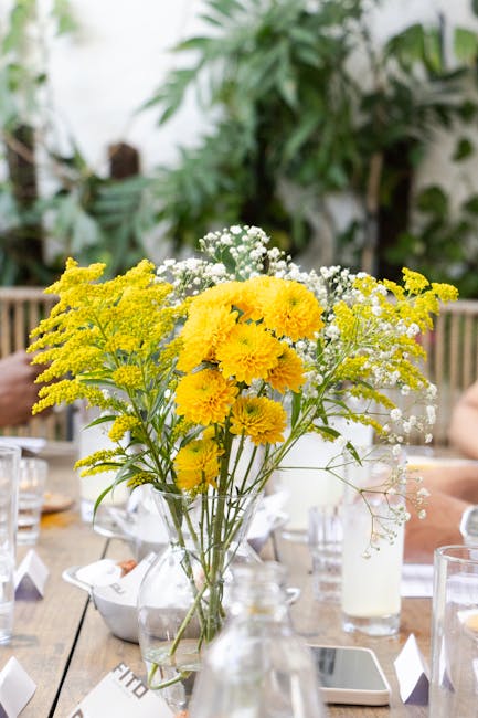 A tall, clear glass vase holding a fresh floral arrangement featuring bright yellow chrysanthemums, delicate white baby's breath, and yellow solidago, set on a wooden table surrounded by glasses, napk