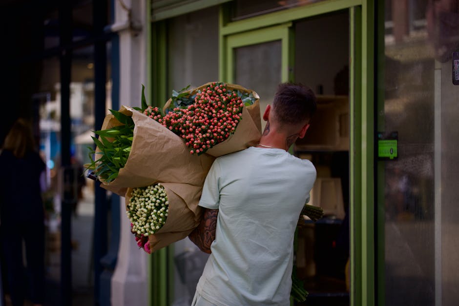 A person with short dark hair, wearing a light grey T-shirt, holds a large bouquet of fresh flowers wrapped in light brown kraft paper, behind their back. The bouquet features pink and white Hypericum