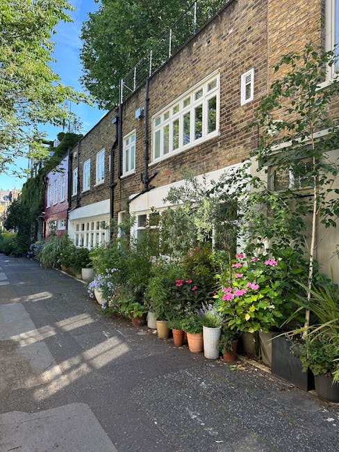 A charming street scene featuring a variety of potted flowering plants arranged along the base of a row of traditional brick houses. The plants include pink, white, and purple blooms with lush green f