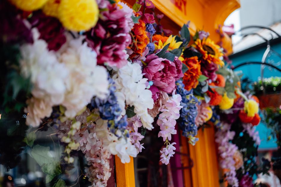 A vibrant floral display featuring an arrangement of fresh flowers, including large pink peonies, white hydrangeas, yellow sunflowers, orange marigolds, and purple delphiniums, all densely clustered t