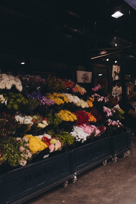A diverse display of fresh flowers arranged on black wheeled carts inside a dimly lit flower shop, with bouquets featuring various colours including yellow, white, pink, red, purple, and green. The fl