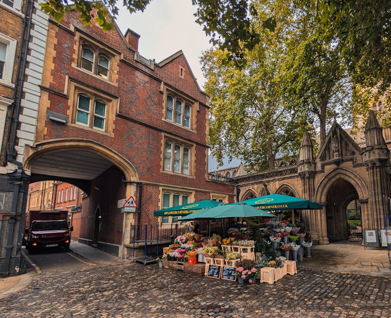 A vibrant outdoor flower stall situated on a cobblestone street beneath a large green umbrella with the signage 'floristsonwellingworth.co.uk.' The stall displays a variety of fresh, colorful flowers 