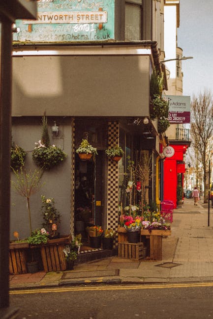 A floral shop on a street corner displays an array of fresh flowers and plants arranged in wooden crates and hanging baskets outside the entrance. The bouquet arrangements feature vibrant colours, inc