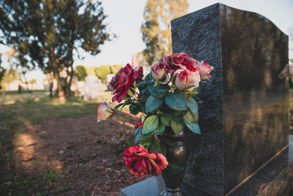 A floral arrangement of pink and red roses, some with white edges, displayed in a black vase positioned next to a polished dark granite headstone in a cemetery. The bouquet appears fresh with vibrant 